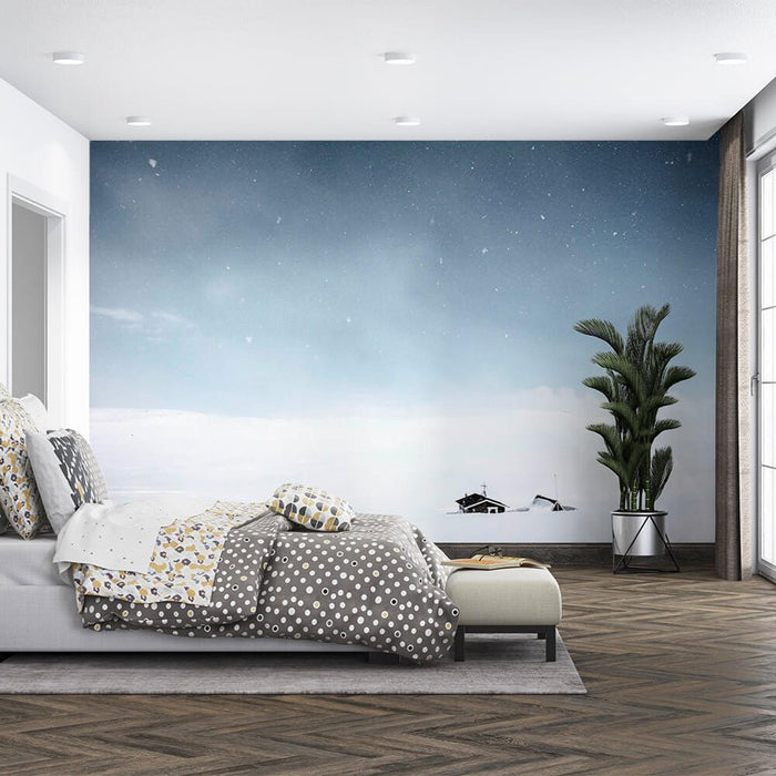 Snowy Wallpaper Mural | Winter Cottage - tranquil snow scene bedroom mural with two small black houses, falling snow, pale blue sky, cozy bedding
